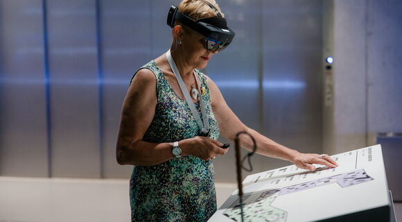 Eine blinde Frau mit kurzen Haaren und Langstock bei der HoloAudioTour im Deutschen Museum Nürnberg. Sie trägt eine AR-Brille, in der einen Hand hält sie eine Fernbedienung. Mit der anderen Hand ertastet sie eine Erklärtafel.