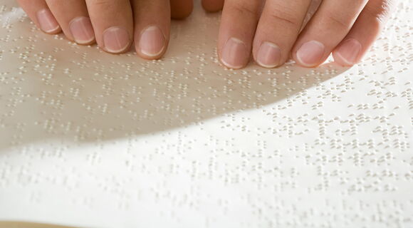 Zwei manikürte Hände lesen Brailleschrift auf weißen Seiten.