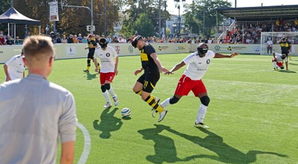 Szene aus einem Blindenfußballspiel: Drei Spieler, davon zwei aus einer Mannschaft, kämpfen um den Ball.