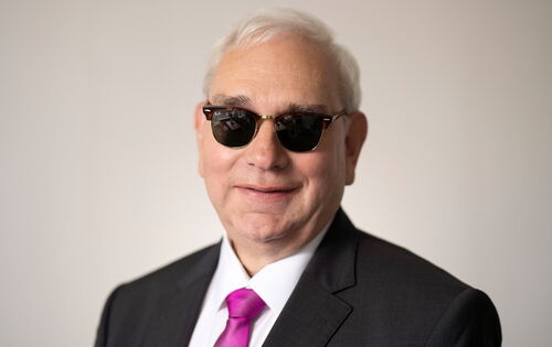 Portrait von Hans-Werner Lange. Er hat kurze, weiße Haare und ein volles Gesicht. Er trägt ein weißes Hemd, darüber ein dunkles Jackett, dazu eine purpurfarbene Krawatte und eine dunkel getönte Brille. Er schaut lächelnd in die Kamera.