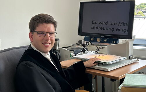 André Stahl sitzt in Richterrobe am Schreibtisch seines Büros. Er hat kurzes dunkleres Haar und trägt eine Brille. Sein Monitor zeigt einen stark vergrößerten Text an.