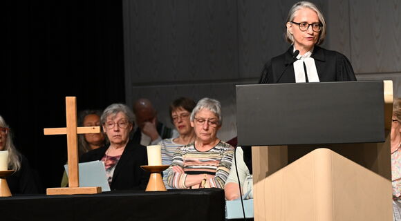 Die Oberkirchenrätin Annette Noller steht im schwarzen Talar am Rednerpult. Sie hat glatte, graue Haare und eine schwarze Brille Auf einem Altar links im Bild stehen ein Holzkreuz und eine weiße Kerze. Dahinter in mehreren Reihen die Mitglieder des Chors.