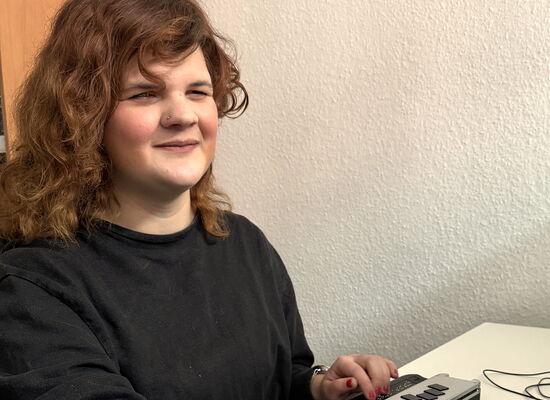 Marie Lampe sitzt an einem Schreibtisch und bedient mit einer Hand die Tastatur, mit der anderen die Braillezeile. Sie hat gewelltes, mittellanges und mitteldunkles Haar.