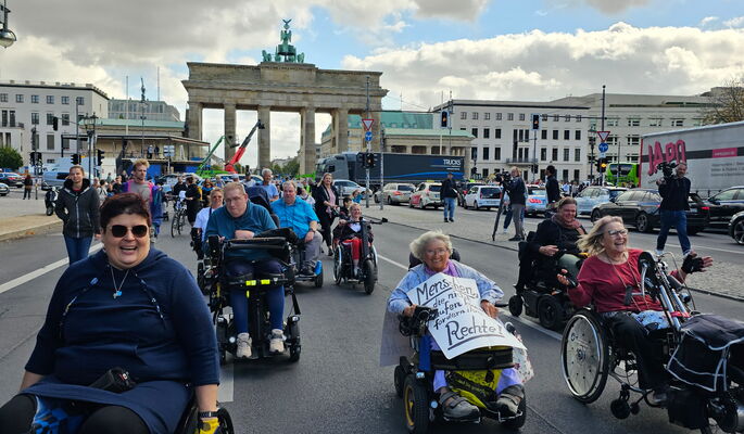 Mehrere Frauen unterschiedlichen Alters fahren strahlend auf der Straße des 17. Juli mit Rollstühlen. Im Hintergrund ist das Brandenburger Tor zu sehen.