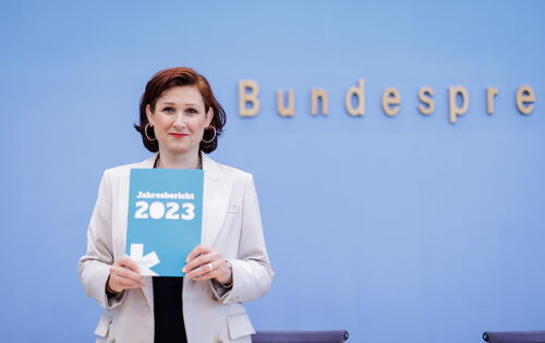 Die Antidiskriminierungsbeauftragte der Bundesregierung, Ferda Ataman, hält den Jahresbericht ihrer Einrichtung in den Händen. Sie hat nackenlanges dunkles Haar, trägt große Kreolen und einen weißen Blazer.