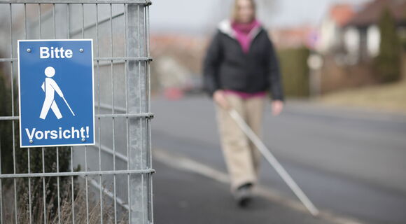 Eine Fußgängerin ist verschwommen im Hintergrund zu sehen. Sie spaziert mit ihrem Langstock auf dem Gehweg. An einem Gitterzaun im Vordergrund hängt ein Schild mit der Aufschrift „Bitte Vorsicht!“ und einem Stockgänger-Symbol.