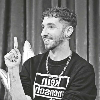 Noam Brusilovsky hebt lachend den linken Zeigefinger. Seine Nägel sind lackiert. Er trägt einen schwarzen Pullover mit dem Aufdruck "Kein Mensch ist illegal".