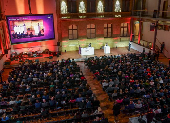 Von schräg hinten wird über das festlich gekleidete Publikum hinweg die Bühne fotografiert. Vorne steht Moderatorin Nadine Heidenreich an einem Pult.