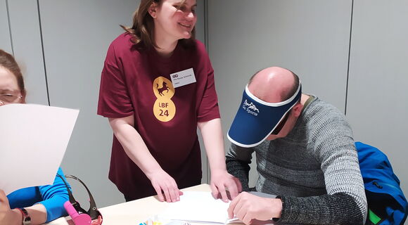 Sonja Schmidt hat schulterlanges, braunes Haar und steht an einem Tisch, auf dem Materialien zum Braillelernen liegen: Eierkartons und Unterlagen.