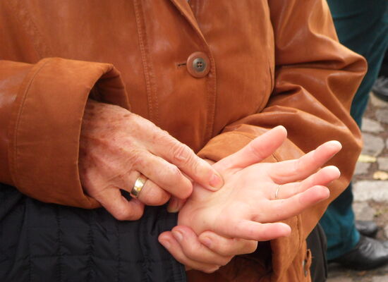 Nahaufnahme von sich berührenden Händen beim Lormen: Die linke Hand einer Person liegt mit der Handfläche nach oben in der linken Hand einer zweiten Person. Diese tippt mit den Fingern ihrer rechten Hand Lormzeichen in die Hand der ersten Person.
