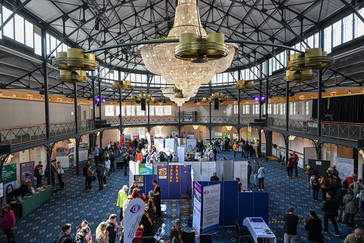 Blick von oben in die Reithalle auf den Markt der Begegnung. Menschen verteilen sich zwischen Ständen, Aufstellern und Fahnen. Der Boden ist blauer Teppich. Fenster rund herum erhellen die Halle. Von der Decke hängen zwei große Kronenleuchter.